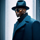 Fashionable man in black overcoat, wide-brimmed hat, and sunglasses poses confidently.