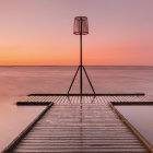 Tranquil sunset over lake with jetty, street lamp, boats, and mountains.