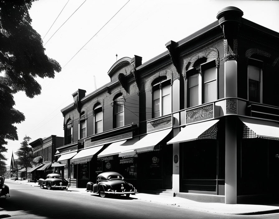 Vintage black-and-white photo of classic 20th-century street with parked cars and two-story buildings.