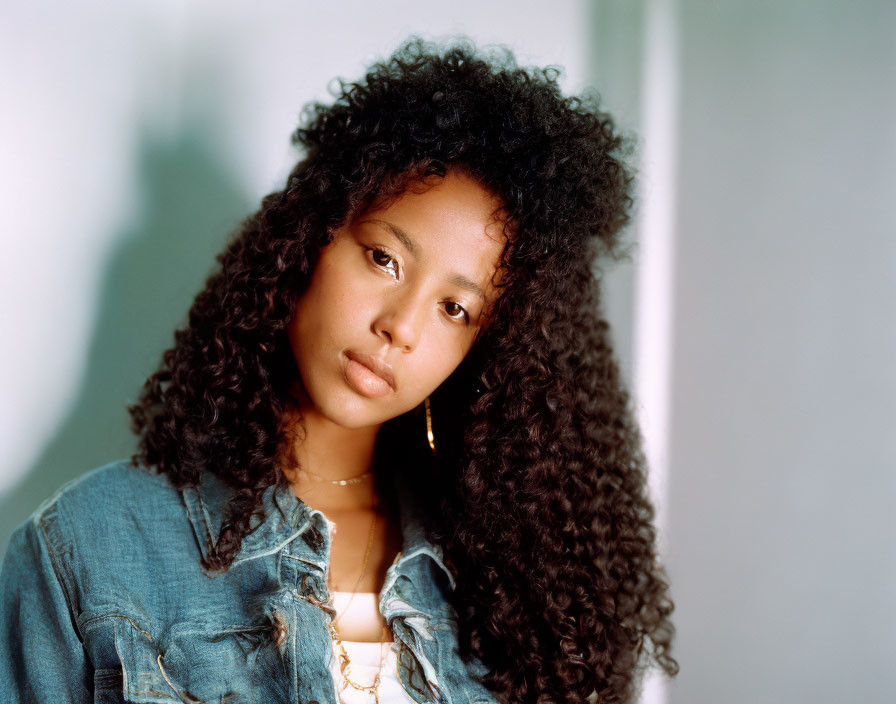 Curly-haired woman in denim jacket with gold earring in soft light.