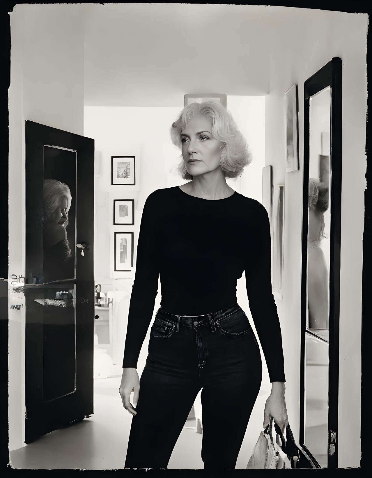 Monochromatic image: Poised woman in black top and jeans, surrounded by framed artworks.