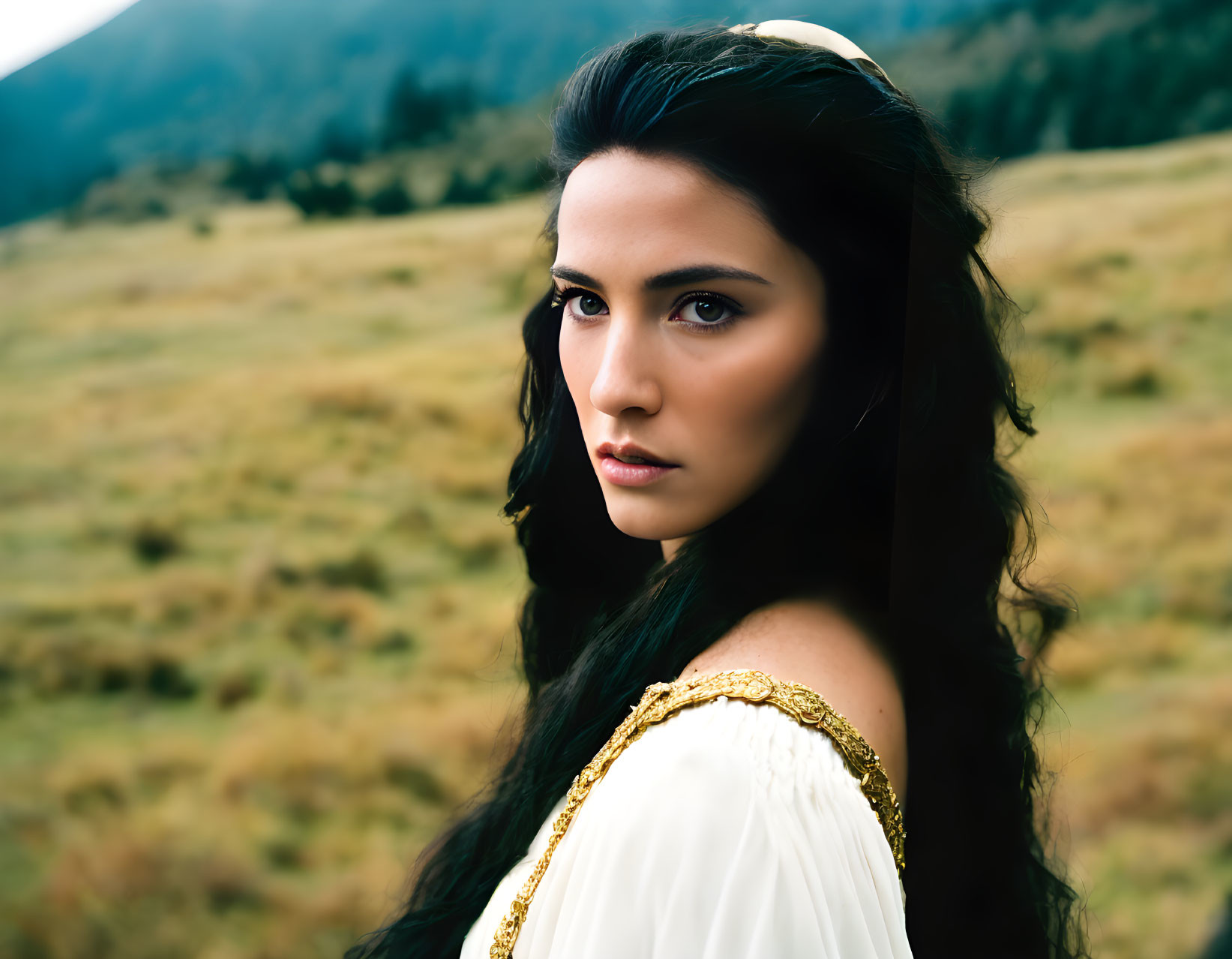 Dark-haired woman in white dress with gold trim in outdoor setting.