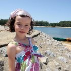 Toddler in pastel dress on sandy shore with canoe and lake.