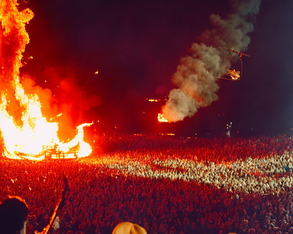 Nighttime fire performance with flaming structure and aerial stunt in front of large crowd