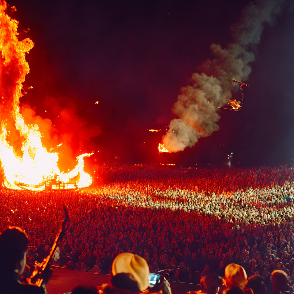 Nighttime fire performance with flaming structure and aerial stunt in front of large crowd