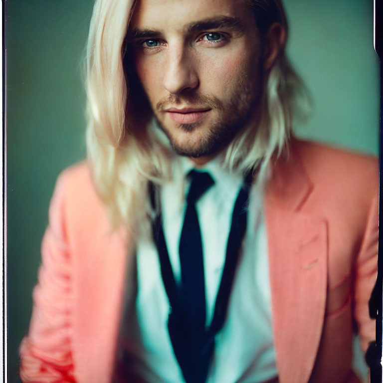 Blonde person in coral blazer, white shirt, navy tie gazes at camera