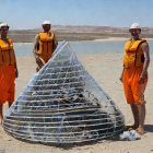 Three Men in Orange Robes with Transparent Cone Structure in Desert