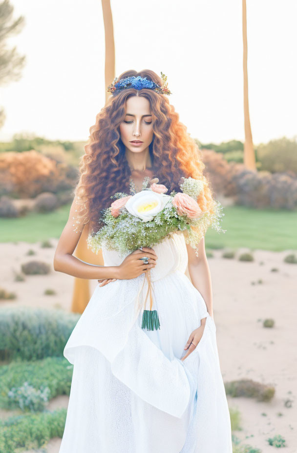 Curly-haired woman in white dress with floral crown holding bouquet outdoors.