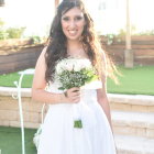 Curly-haired woman in white dress with floral crown holding bouquet outdoors.