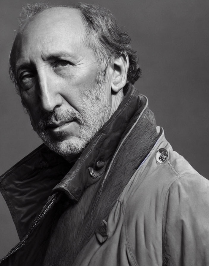 Monochrome portrait of mature man with beard in jacket and emblem, looking thoughtful.