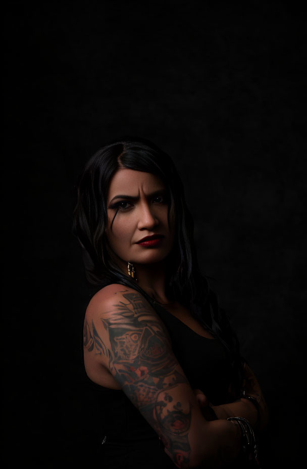 Dark-haired woman with tattoos and striking makeup gazes at camera on black backdrop