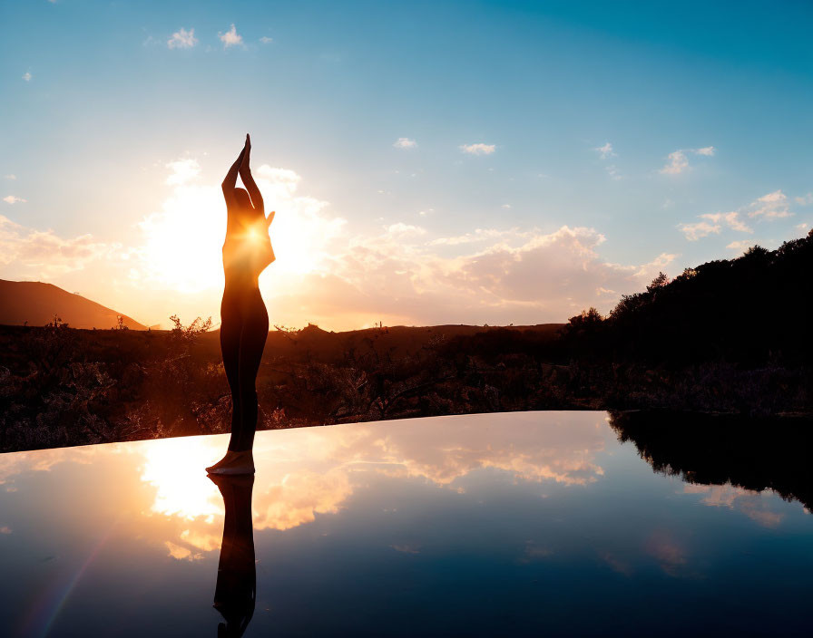 Silhouette of person in yoga pose at sunrise or sunset with reflective surface and serene landscape.