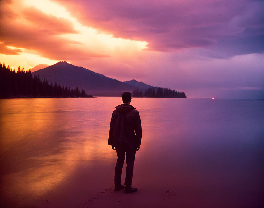 Tranquil sunset scene: person on shore, purple and orange sky, calm water, distant mountains