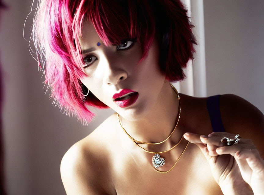 Vibrant woman with pink hair and bold makeup holding an object in gold necklace portrait.