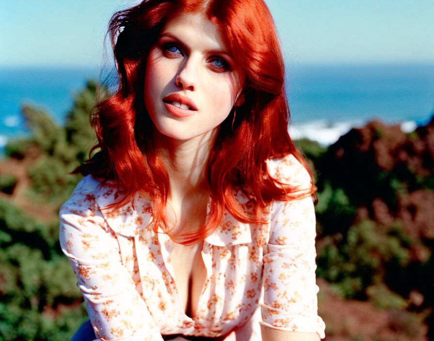 Red-haired woman in floral blouse outdoors with trees and blue sky.