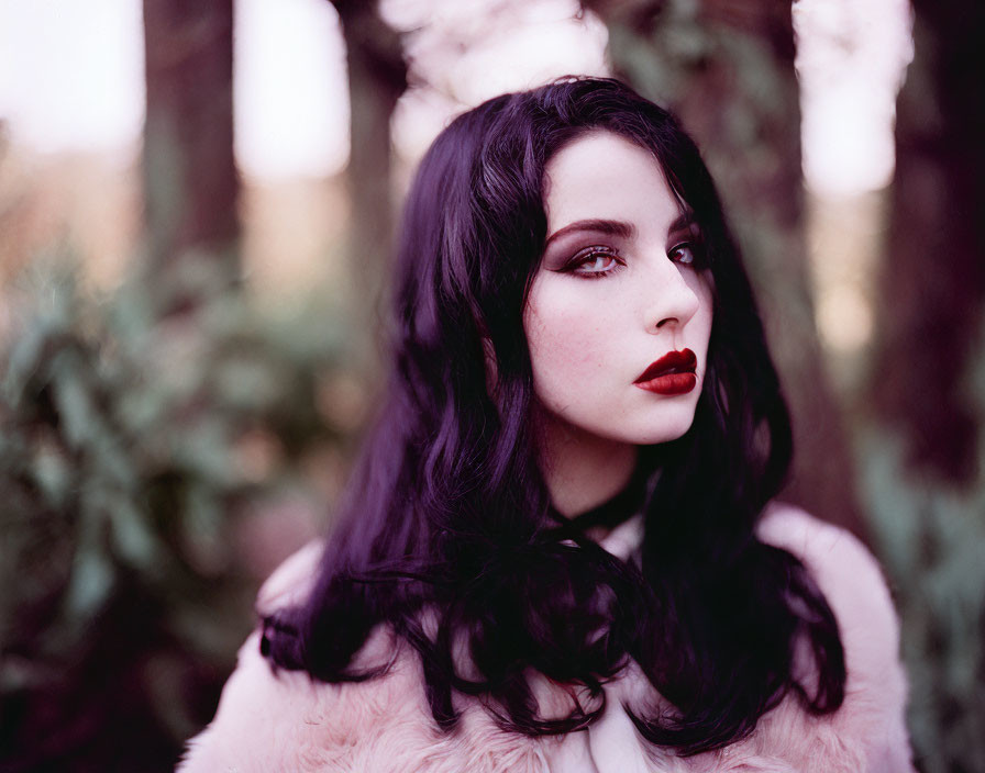 Dark-haired woman in red lipstick wears pink fur coat outdoors with dreamy forest backdrop