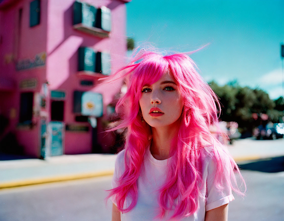 Pink-haired woman with matching lipstick in front of pink building under blue sky