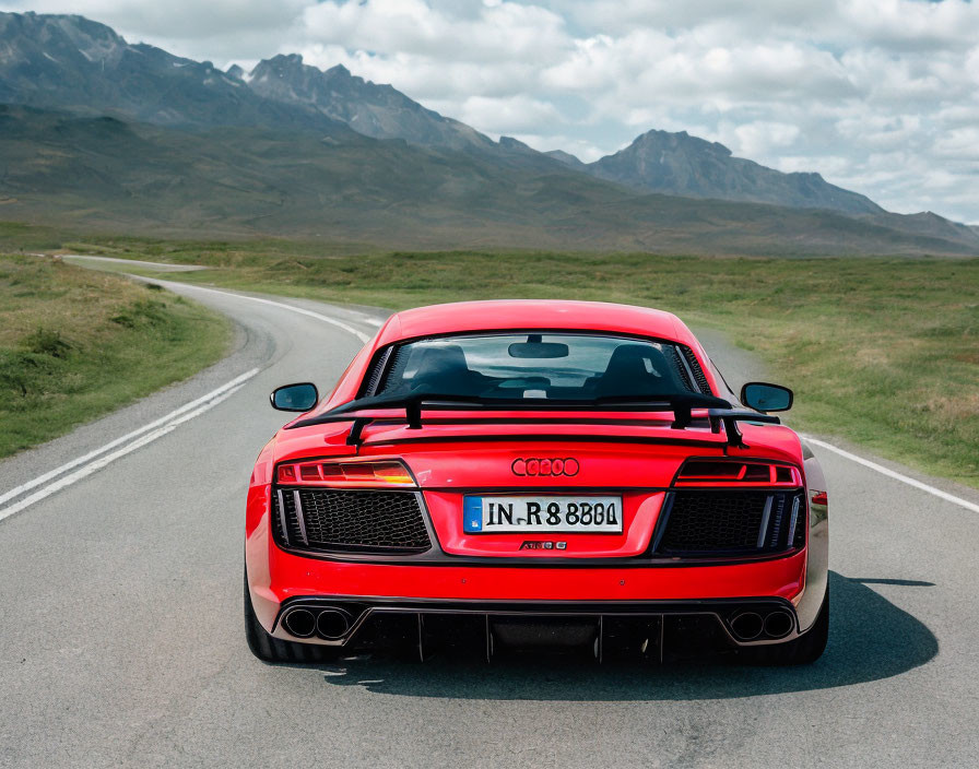 Red Audi R8 sports car on winding road in grassy landscape with mountains and cloudy sky.