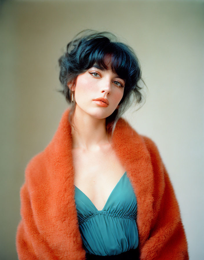 Dark-haired woman in orange fluffy coat and teal dress gazes softly.