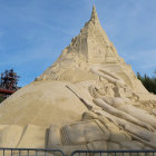 Detailed sandcastle with tiers and turrets against blue sky and buildings.