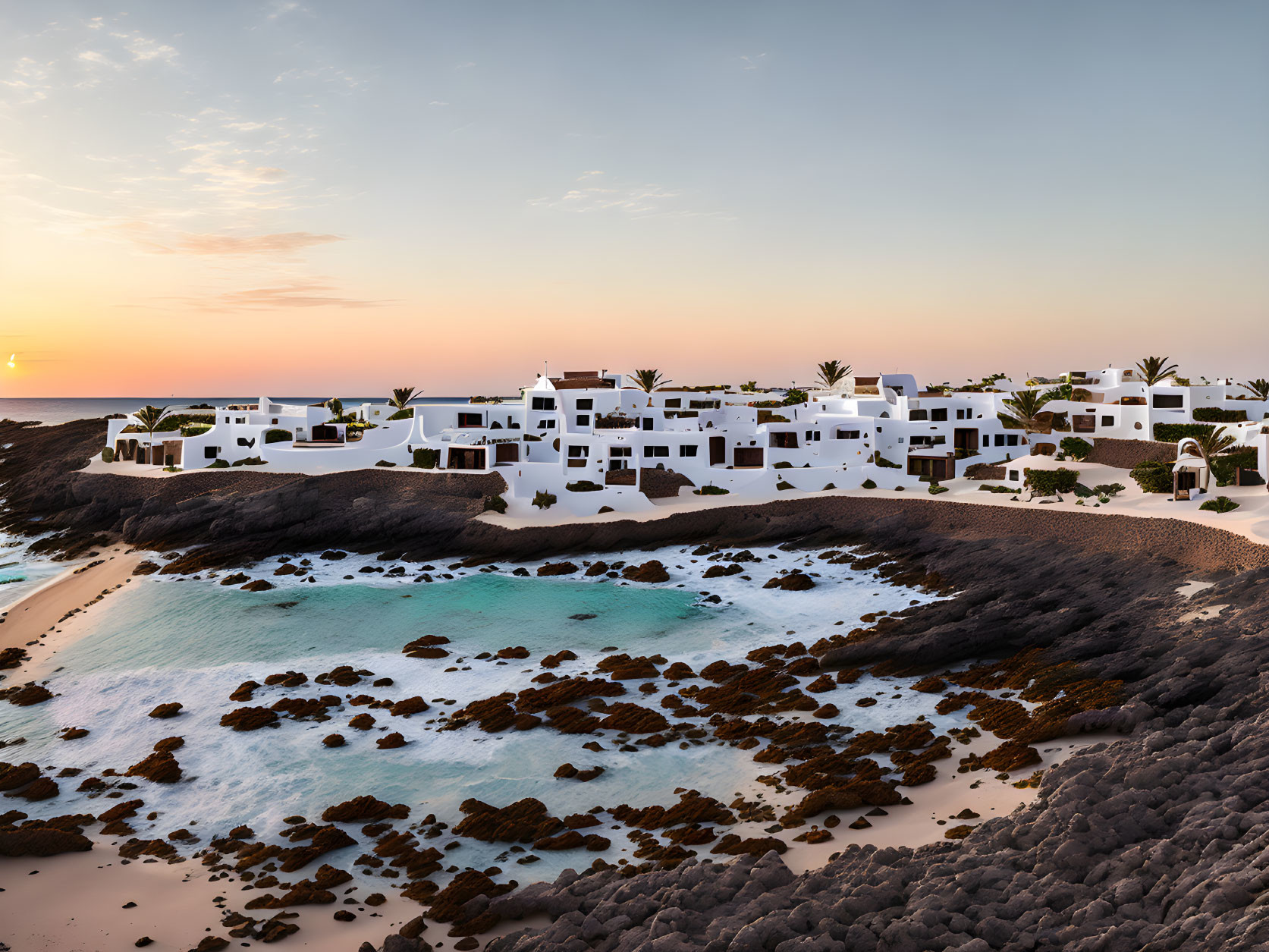 Coastal sunset with white buildings, sandy beach, clear blue water, and rocky outcrops