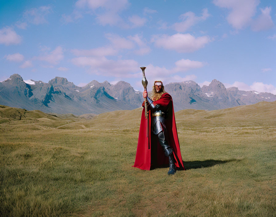 Armored figure with red cape in grassy field with mountain range.