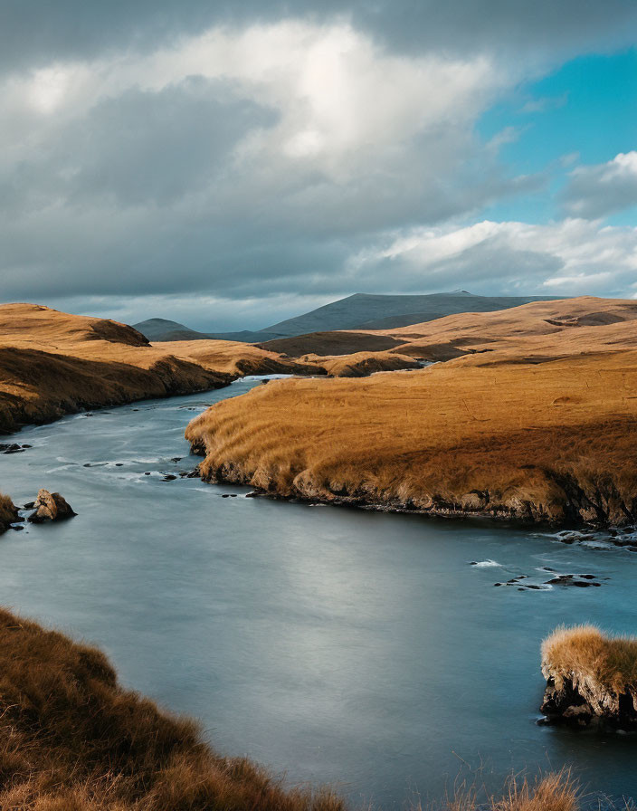 Scenic river winding through hilly landscape with golden grass under dramatic sky