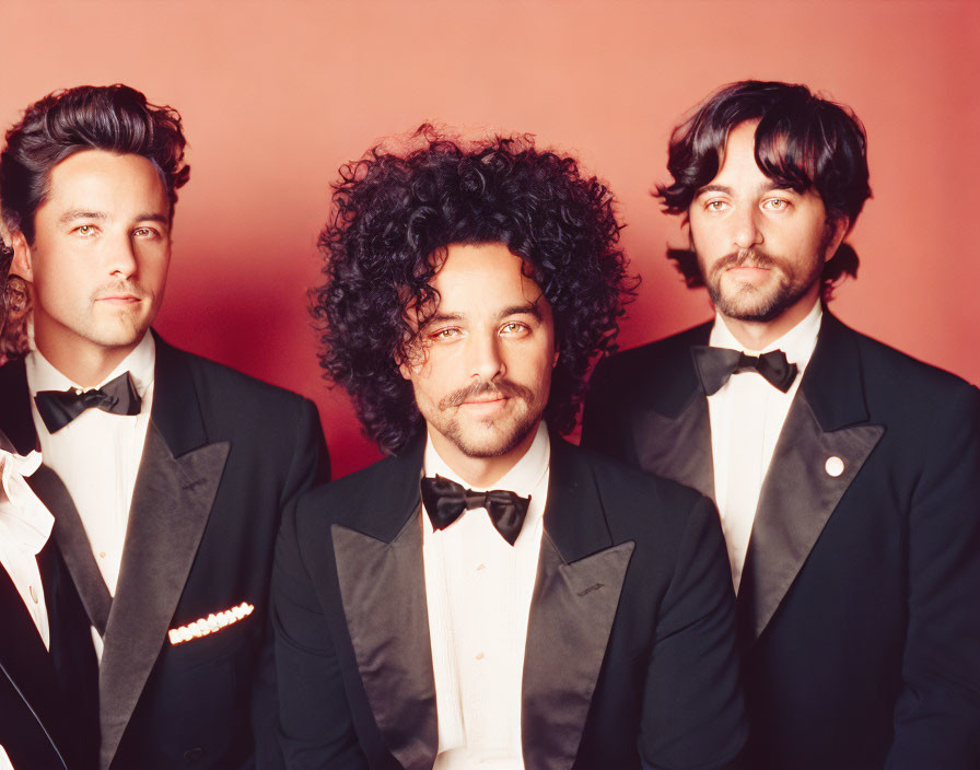 Three men in tuxedos with bow ties against red background; central figure with curly hair.