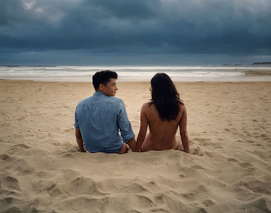 Couple sitting on beach, facing stormy ocean skies