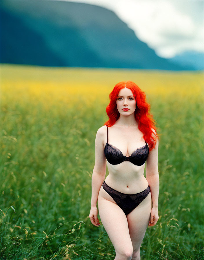 Red-haired woman in black lingerie standing in yellow field with mountains.