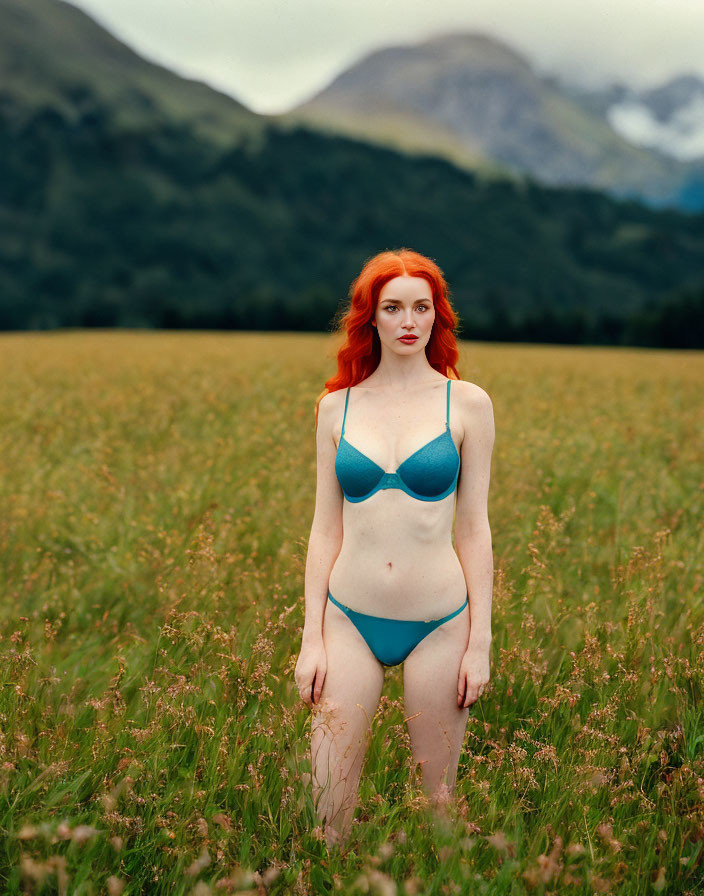 Vibrant red-haired woman in teal bikini in field with mountains & cloudy sky