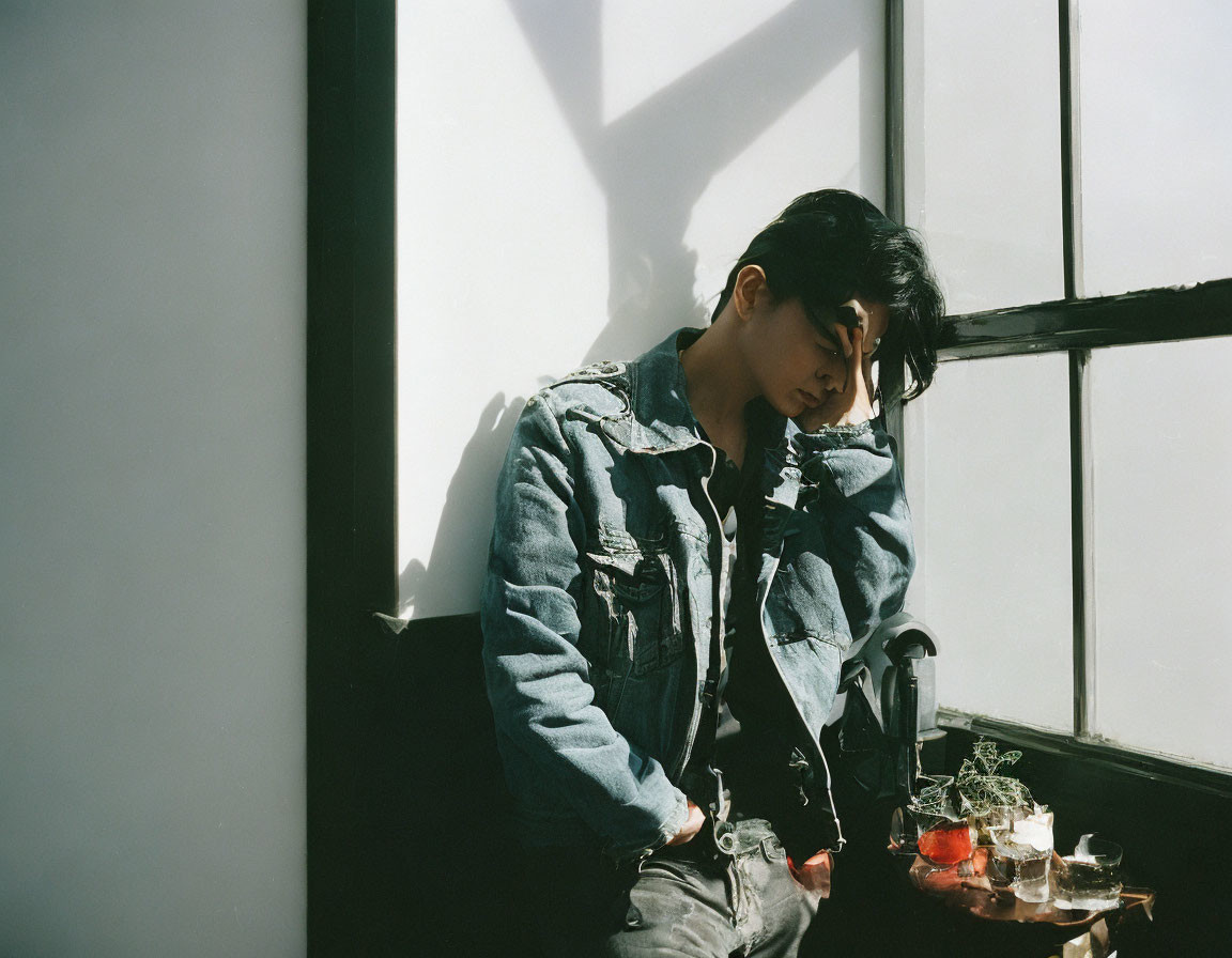 Young man in denim attire sitting by window with pensive expression and sunlight shadows.