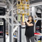 Red-haired woman in black clothing poses confidently with complex quantum computer setup