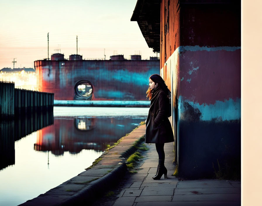 Woman in black coat by canal at sunset with industrial backdrop