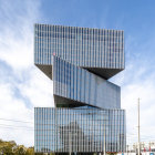 Contemporary glass building with undulating facade, trees, water feature, and birds under blue sky