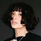 Portrait of a person with curly black hair, round glasses, freckles, and dark garment.