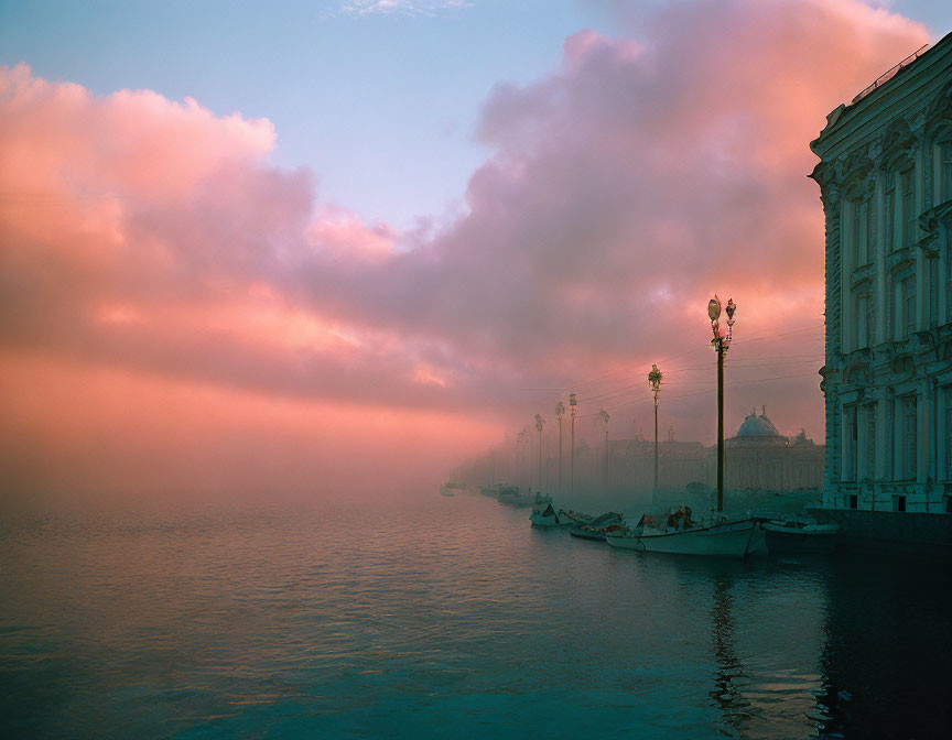 Tranquil riverscape at dawn or dusk with pink and blue sky hues, street lamps, and