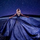 Woman in flowing blue gown gazes at distant mountain under starry night sky