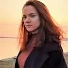 Profile of young woman with wavy hair at sunset by water in dark coat and pink top