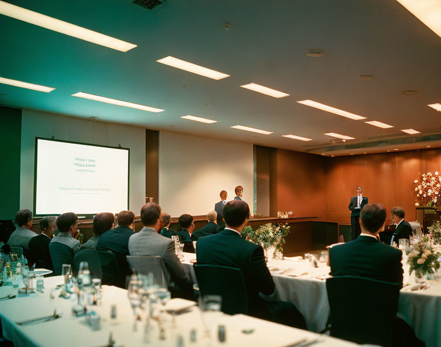 Conference room business presentation with attendees seated at tables facing speaker and screen slide.