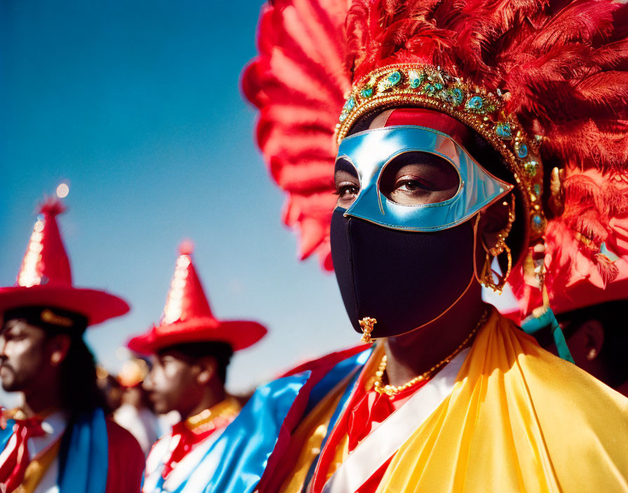 Person in blue mask and elaborate carnival attire with red feathers, among others in colorful costumes under clear sky