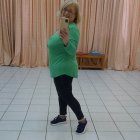 Blonde person in green dress and black leggings posing against curtain backdrop