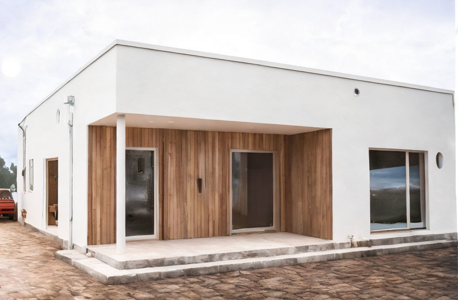 White-walled single-story house with wooden door, large windows, paved front yard, cloudy sky