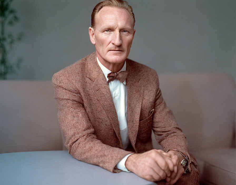 Fair-skinned man with ginger hair in brown suit and bow tie on couch