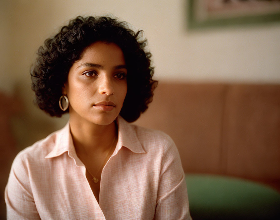 Curly-haired woman in pink striped shirt and hoop earrings sitting indoors