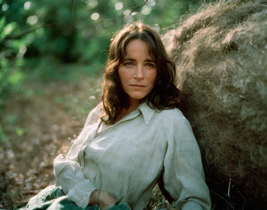 Woman with medium-length hair by hay bale in forest, wearing light striped shirt