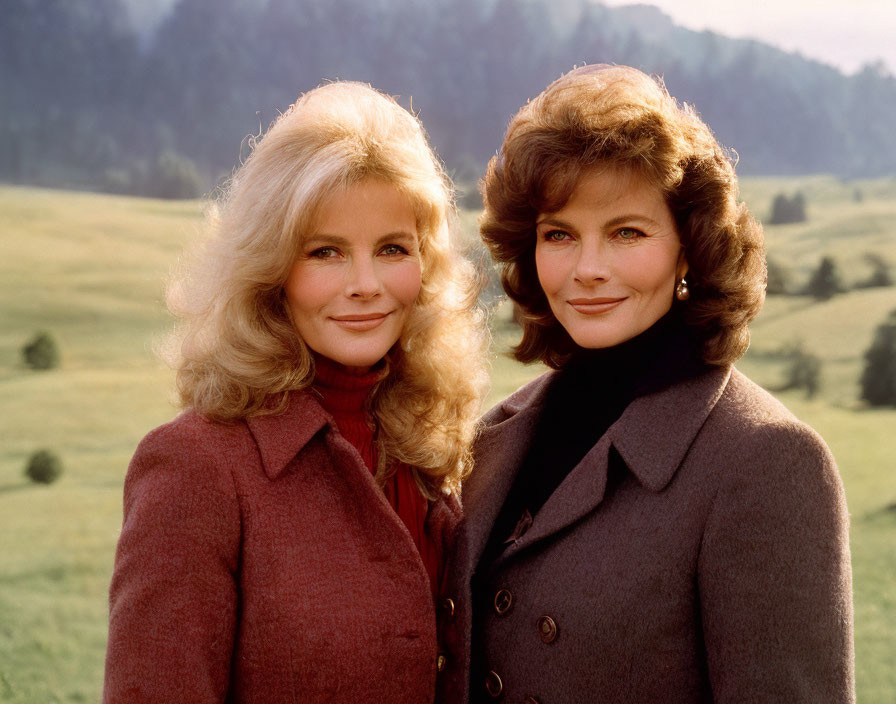 Two women with voluminous hairstyles in elegant coats posing outdoors with green hills backdrop