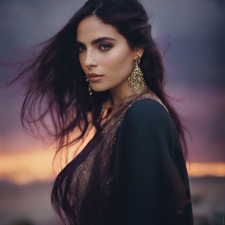 Dark-haired woman in gold earrings wearing black garment against dusky sky