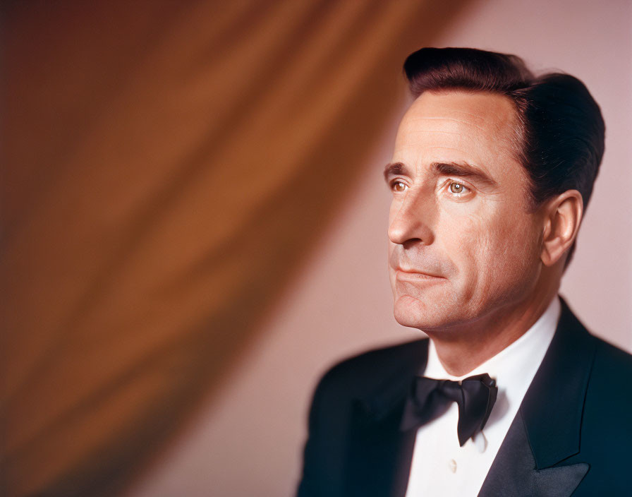 Formal man in tuxedo with bowtie and slicked-back hair in thoughtful pose on warm