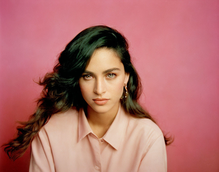 Woman with wavy hair in pink shirt against pink backdrop gazes at camera.